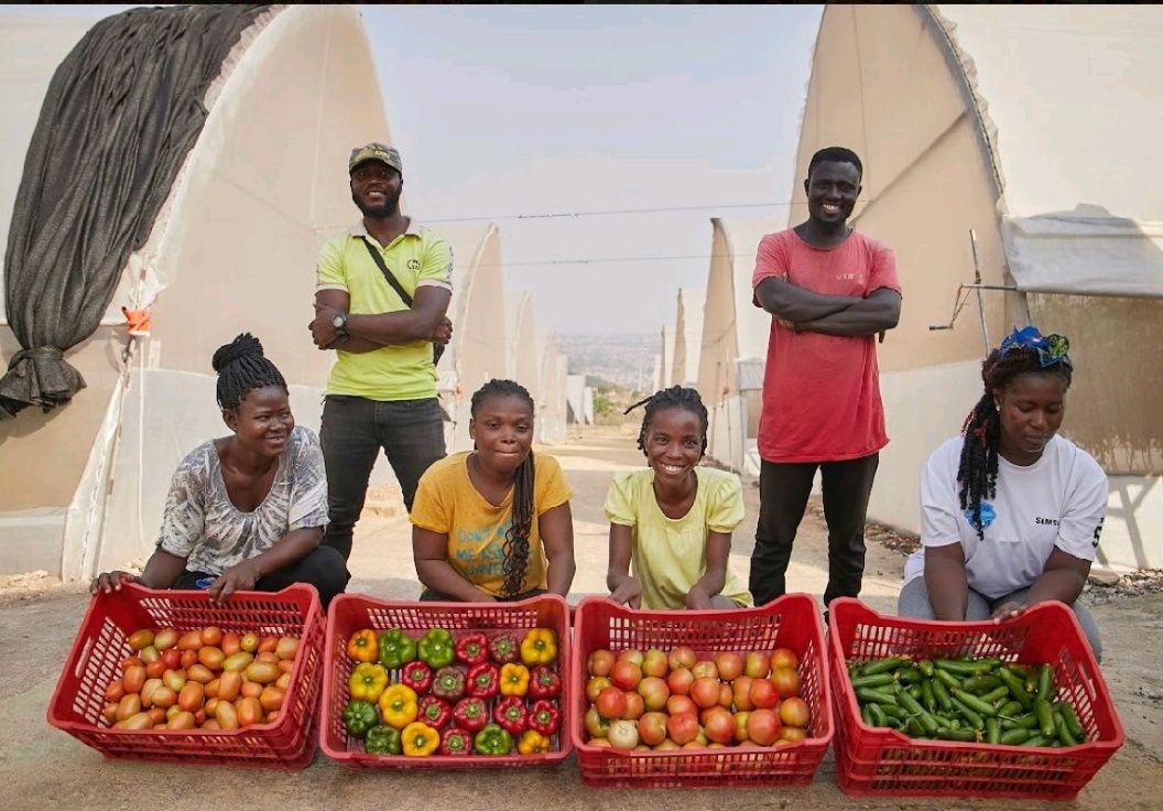 Farm workers smiling with baskets of fresh vegetables in front of greenhouses.