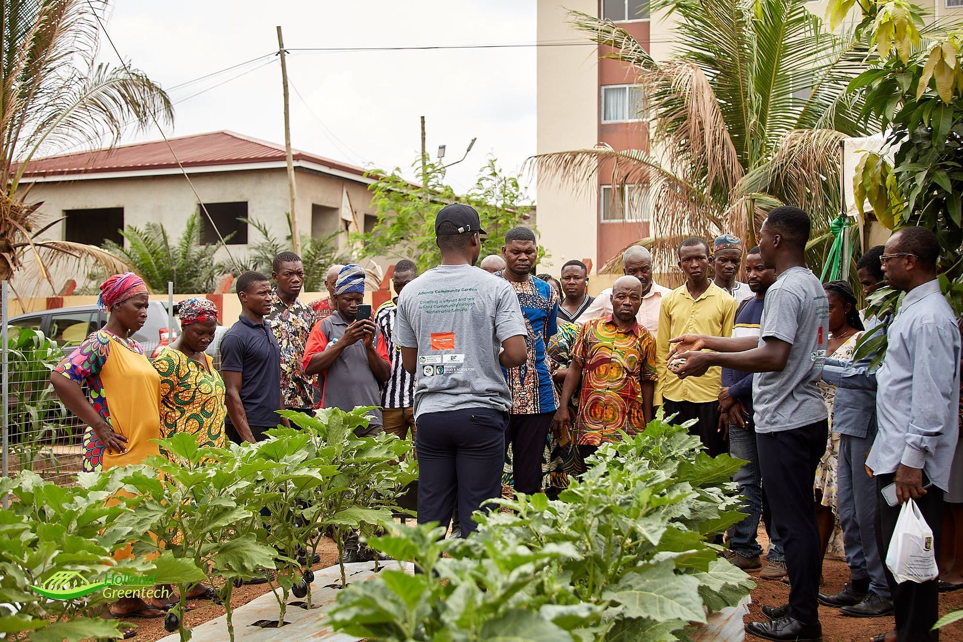 Group of people gathered around plants in a community garden, receiving a presentation.